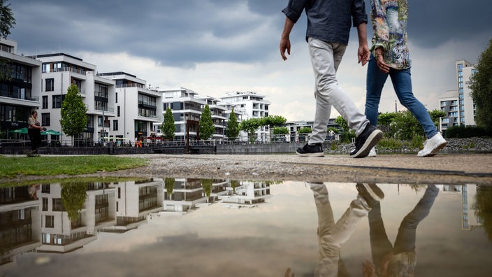 Zwei Spaziergänger spiegeln sich unter dunklen Wolken am Phoenix See in einer Pfütze. Zum Start ins Wochenende werden in Nordrhein-Westfalen Schauer und Gewitter erwartet. 