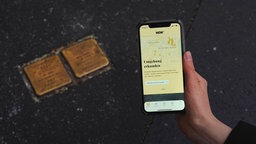 Eine Hand hält ein Smartphone mit der App "Stolpersteine NRW" über zwei Stolpersteine auf dem Gehweg