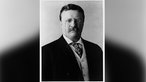 Offizielles Porträt von US-Präsident Theodore Roosevelt: Schwarz-weißes Bild eines Mannes mit einer Brille und einem Schnauzbart in Anzug mit Weste, der ernst in die Kamera schaut