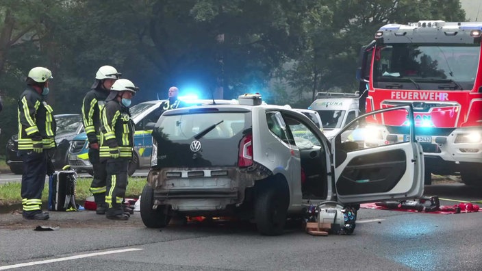 Feuerfehrpersonal steht neben dem zerstörten Unfallfahrzeug. Im Hintergrund Feuerwehr- und Polizeiautos