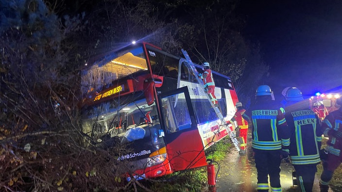 Ein Bus ist in einen Straßengraben gefahren und wird von einem Feuerwehrmann auf einer Leiter bestiegen. 