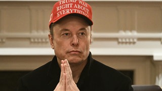 Elon Musk sitzt mit roter Kappe auf einem Stuhl.