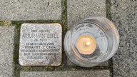 Links der Stolperstein von Artur Manteufel, rechts daneben ein Teelicht im Glas