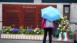 Gedenkstätte mit bunten Blumen und Kerzen. Davor steht eine Person mit rosa Jacke und einem hellblauen Regenschirm