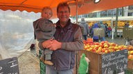 Norbert Weber vor seinem Marktstand und Kind auf dem Arm
