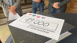 Auf einem Tisch liegt ein Blatt mit der Aufschrifft "19.000 Unterzeichnerinnen und Unterzeichner"