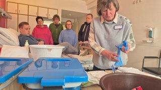 Eine Frau steht am Tisch mit den Krankenhausutensilien, im Hintergrund sieht man die Kursmitglieder.