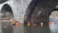 Zwei Männer in Tauchausrüstung arbeiten im Wasser am Fuß eines Brückenpfeilers.