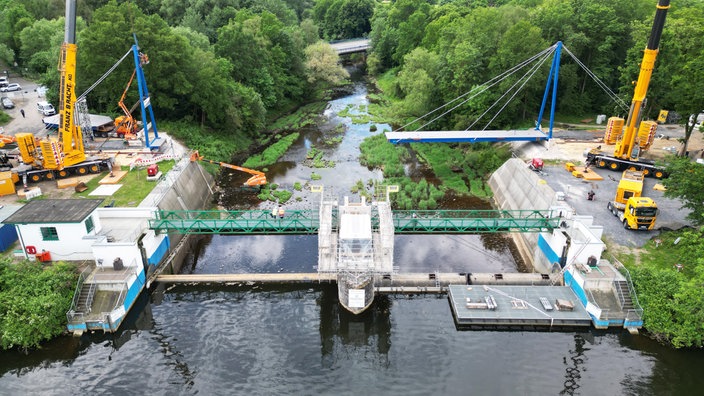 Hinter einem Staudamm ist eine Baustelle, auf der eine Brücke gebaut wird.