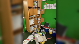 Auf dem Bild ist ein Holzschrank mit verschiedenen Fächern zu sehen. Die Türen sind aufgerissen. Akten, Papiere und Hefter liegen herausgerissen auf dem Boden.