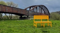 Zu sehen ist eine gelbe Bank, welche auf einer grünen Wiese steht. Hinter ihr verläuft eine alte Brücke aus Metall in einem rostrot.