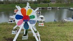 Zu sehen ist ein Miniaturriesenrad, welches bunt bemalt ist. Es steht auf einer Wiese vor einem See.