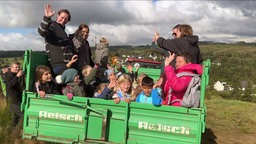 Viele Kinder mit Erwachsenen sitzen auf einer Ladefläche von einem Trecker und winken.