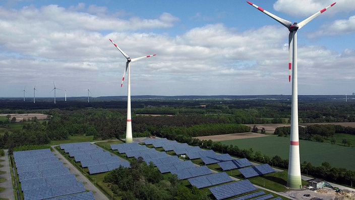 Zwei Windräder stehen auf einer freien Fläche, darunter Solaranlagen.