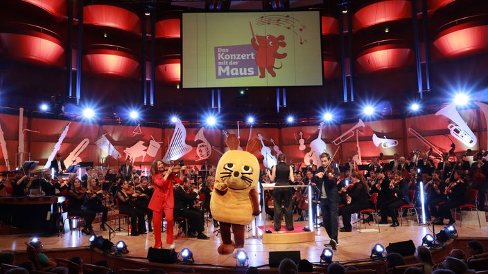 Die Maus, Jana Forkel und Johannes Büchs stehen gemeinsam mit Musiker*innen des WDR Sinfonieorchesters auf der Bühne der Kölner Philharmonie.