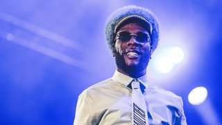 Burna Boy, mit einem Weißen Hemd, Fellkappe, Sonnenbrille  und Schlips auf der Bühne. 