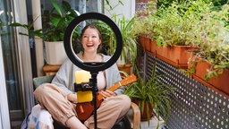 Blick durch ein Ringlicht mit daran montiertem Smartphone: Ein junge Frau sitzt lachend mit einer Ukulele auf einem Balkon