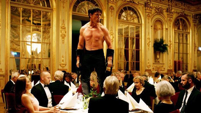 Filmszene aus "The Square". Ein halbnackter Mann steht auf einem Tisch in einem Nobelrestaurant