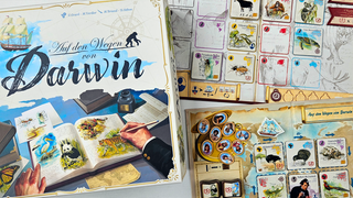 Die Spielschachtel von "Auf den Wegen von Darwin", daneben die zentrale Auslage und das Reisetagebuch eines Spielers, auf denen bunte Tierplättchen liegen