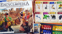 Die Spieleschachtel von "Encyclopedia" neben dem farbenfrohen Spielplan, auf dem Karten mit bunten Tierillustrationen und Würfel liegen