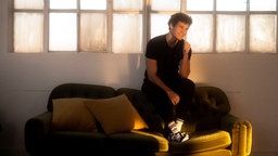 Wincent Weiss sitzt in schwarzem Outfit auf einer grünen Couch die von etwas Sonnenlicht angestrahlt wird.