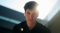 Eien Nahaufnahme von Wincent Weiss vor einem verschwommenen Gebäude und einem strahlenden Himmel.