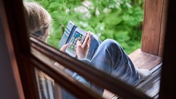 Ein Kind sitzt am Fenster und schaut auf sein Handy.