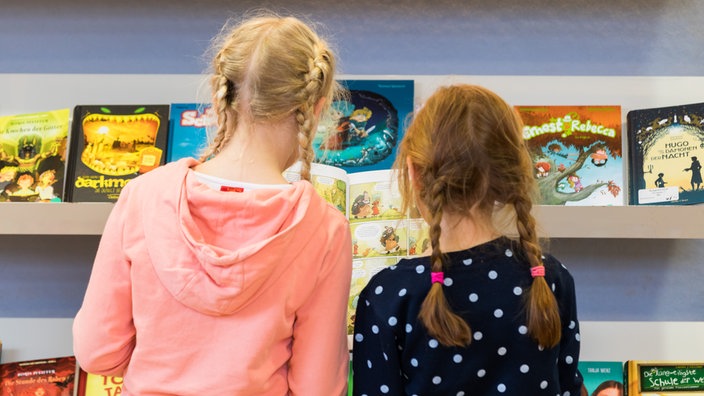 Zwei Kinder stehen mit dem Rücken zur Kamera und schauen sich gemeinsam ein Buch an, an der Wand ein Ausstellungsregal mit Büchern.