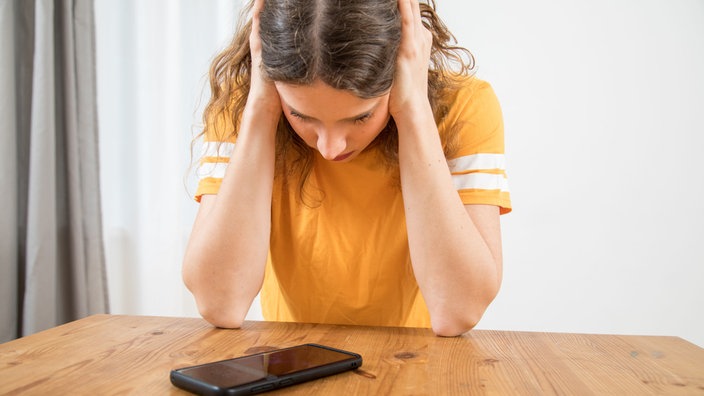 Eine junge Frau sitzt an einem Tisch und sich die Ohren zu während sie auf ihr Handy schaut.