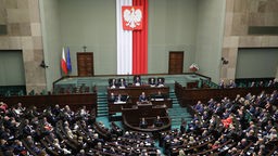 Aufnahme des polnnisches Parlamentes bei einer Abstimmung, hinter dem Rednerpult eine große Flagge in den rot-weißen Nationalfarben Polens.