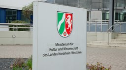 Außenansicht des Geländes sowie ein Hinweisschild mit dem Schriftzug "Ministerium für Kultur und Wissenschaft des Landes Nordrhein-Westfalen" und Landeswappen.