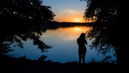 Die Silhoutte eines Menschen an einem bewaldeten See, in dem sich die untergehende Sonne spiegelt.