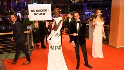 Model Papis Loveday mit einem Schild "No Racism No AfD" neben Nikolai Kinski auf dem Roten Teppich bei der Eroeffnung der Berlinale. 