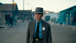 Filmszene: Cillian Murphy als Oppenheimer, gekleidet im Stil der 1940er Jahre, auf einer staubigen Straße zwischen kleinen Häusern.