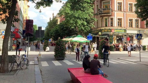 Symbolbild: Eine autofreie Straße in Stockholm im Jahr 2017 mit Passanten, Radfahrenden, Sitzgelegenheiten und Grünpflanzen auf der Fahrbahn.