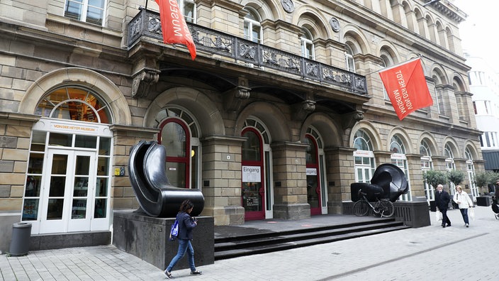 Fahnen wehen am von der Heydt-Museum. Menschen gehen auf dem Bürgersteig daran vorbei.