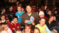 Jan Josef Liefers mit einem Buch in der Hand umgeben von Kindern