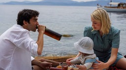 Leonard (gespielt von Alex Wolff) sitzt mit Marianne Ihlen (gespielt von Lise Fjeldstad) und einem Kleinkind in einem Boot.