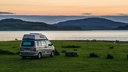 Im Abendlicht steht ein Campervan auf einer grünen Wiese zwischen Schafen direkt an einem idyllischen Platz am Meer. Grossbritannien.