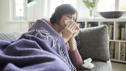 Frau liegt auf einem Sofa unter einer Decke und putzt sich die Nase