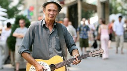 Abi Wallenstein, in einer Fußgängerzone mit Gitarre, im Hintergrund unscharf einige Passanten