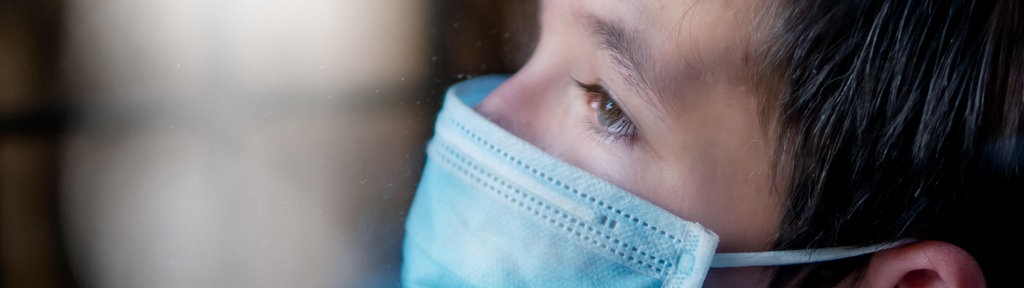 Trauriger Junge mit medizinischer Gesichtsmaske hinter einem Fenster während der Ausganssperre