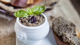 Tapenade serviert in einer kleinen Schüssel. Daneben ein mit Tapenade bestrichenes Baguette.