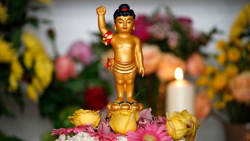 Buddhistisches Vesak-Fest: Eine kleine goldene Figur steht inmitten von Blumen, im Hintergrund brennt eine Kerze.