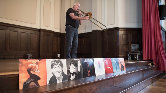 Eduard Meyer mit Posaune: Im Vordergrund stehen diverse, aufgereihte LPs seiner Schaffenszeit als Tontechniker, ua. von David Bowie.