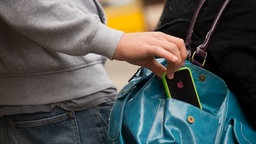 Symbolbild: Diebstahl und Ursünde. Eine Hand greift in die Handtasche einer Frau und entwendet ein Smartphone mit einem Apfel darauf.