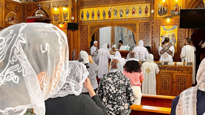 Das Bild zeigt eine koptische Messe im Kloster St.-Antonius. Mitglieder der Gemeinde stehen dabei in einem Kirchenraum mit hölzerner Vertäfelung vor einem Altar.