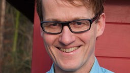 Das Bild zeigt Björn Süfke, einen Psychologen. Der Mann hat kurze braune Haare und trägt eine schwarze Brille, er trägt dazu ein blaues Hemd und lächelt.