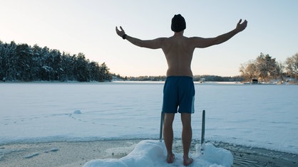 Mann mit Badehose und Wintermütze steht mit ausgebreiteten Armen vor einem Eisloch mit Leiter