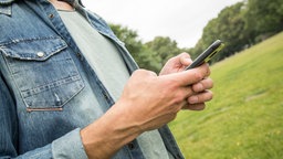 Junger Mann hält Smartphone in der Hand, im Hintergrund Bäume